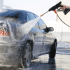 Ruční vysokotlaká myčka čistí tmavě modrý automobil za zimního počasí, přičemž proud vody stříká na karoserii vozu.