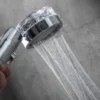 Detail sprchové hlavice s proudem tekoucí vody, zachycené z blízka na šedém pozadí.