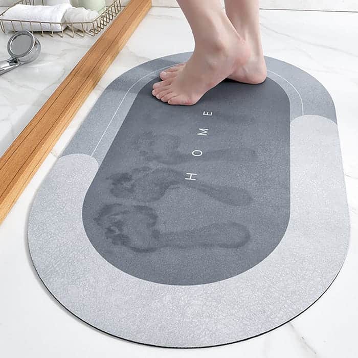 Absorpční šedá koupelnová rohož s designem Home, na které jsou viditelné šlápoty nohou, umístěná v koupelně vedle dřevěné lišty.