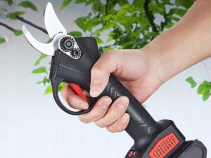 Elektrické zahradní nůžky s černým tělem a kovovými ostřími, které drží ruka na zelené listnaté pozadí.