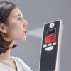 A person in profile blowing towards a digital breath alcohol tester device, which displays readings on its LCD screen against a neutral gray background.