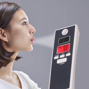 A person in profile blowing towards a digital breath alcohol tester device, which displays readings on its LCD screen against a neutral gray background.