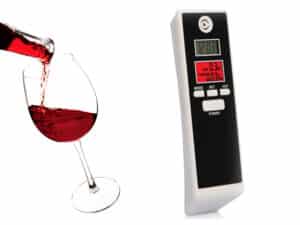 Digitálny alkohol tester s displejom vedľa pohára červeného vína, slúžiaci na rýchle a jednoduché zmeranie hladiny alkoholu v dychu pred šoférovaním.