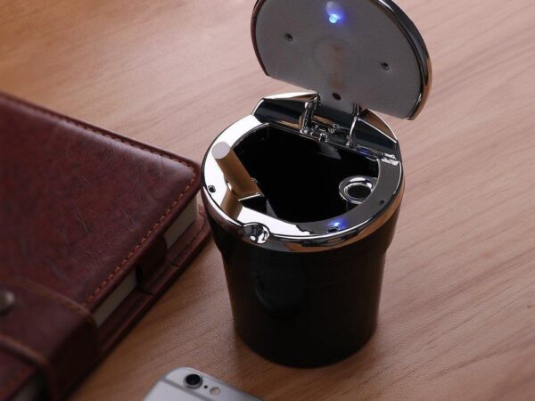 Černý kovový popelník s kovovým víkem a modrým LED osvětlením, umístěný vedle hnědé kožené peněženky a mobilního telefonu na dřevěném povrchu.