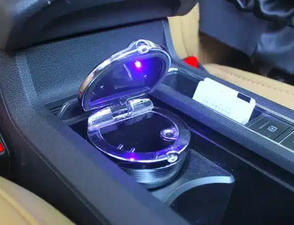 Modrým LED světlem osvětěný kovový popelník v konzoli automobilu, částečně otevřený.