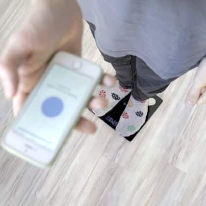Pohled shora na osobu v ponožkách s motivem a mobilní telefon s rozmazaným modrým displejem, položené na světlé podlaze.
