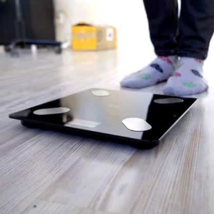 Digitální osobní váha stojící na dřevěné podlaze, vedle které jsou vidět nohy v ponožkách s puntíky.