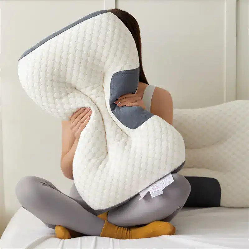 Ergonomický bílo-šedý masážní polštář ve tvaru asymetrického tvaru, který někdo drží v náručí a sedí na posteli v pohodlné póze.