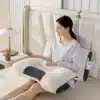 Ergonomický bílý masážní polštář s černými detaily, na kterém sedí žena v bílém oblečení a odpočívá na světlém pohovce vedle nočního stolku s lampou.