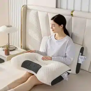 Ergonomický bílý masážní polštář s černými detaily, na kterém sedí žena v bílém oblečení a odpočívá na světlém pohovce vedle nočního stolku s lampou.