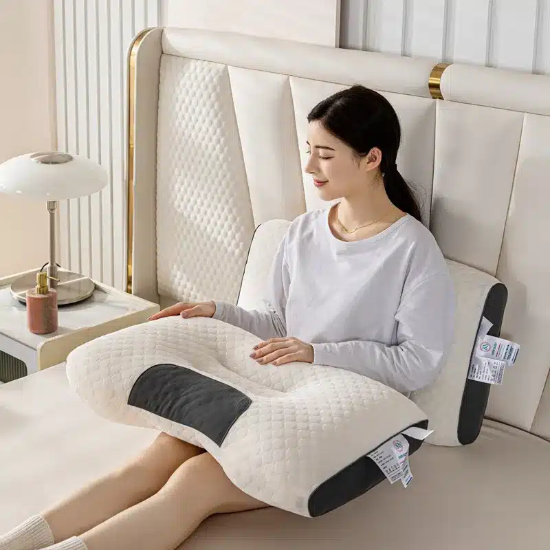 Ergonomický bílý masážní polštář s černými detaily, na kterém sedí žena v bílém oblečení a odpočívá na světlém pohovce vedle nočního stolku s lampou.