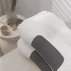 Ergonomický masážní polštář v bílé a tmavě šedé barvě umístěný na bílém ručníku vedle dekorativní misky na minimalistickém pozadí.
