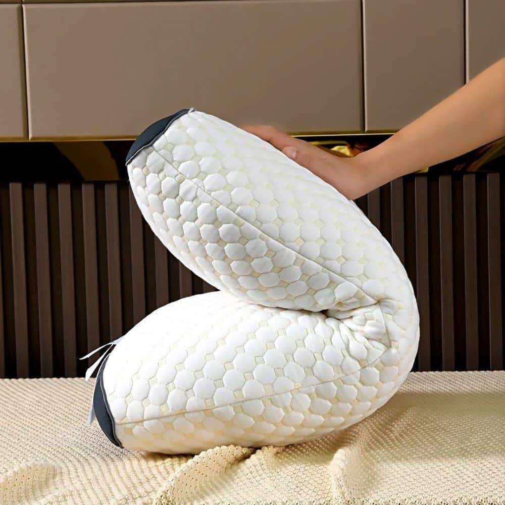 Bílý ergonomický masážní polštář s hexagonálním vzorem, který je tvarován do zakřiveného tvaru a ležící na béžovém koberci vedle tmavě hnědé stěny, s rukou dotýkající se jeho horní části.