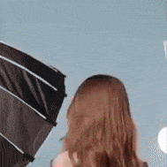 Zadní pohled na ženu s hnědými vlasy, která je otočená k šedému pozadí, vedle ní je částečně viditelný tmavý předmět.