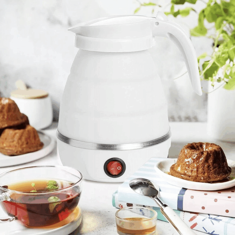 Bílá elektrická konvice na stolku s šálkem čaje, sladkým pečivem a čajovým lístkem v pozadí, připravená k vaření vody.
