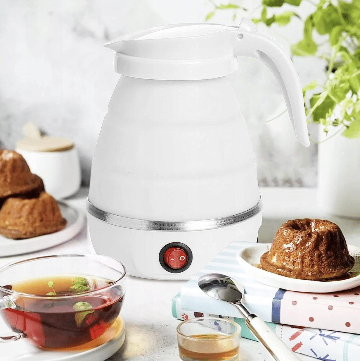 Bílá elektrická konvice na stolku s šálkem čaje, sladkým pečivem a čajovým lístkem v pozadí, připravená k vaření vody.