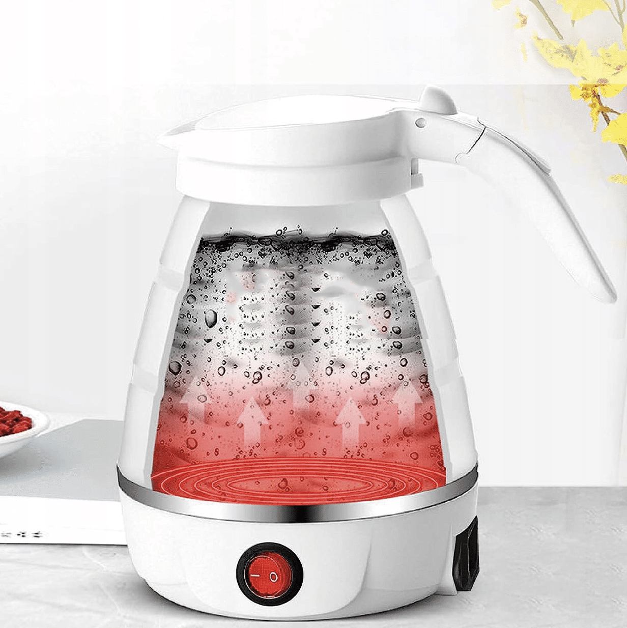 Elektrická skleněná konvice s červeným osvětlením dna, naplněná vodou se vzduchových bublinkami, umístěná na bílém podstavci s vypínačem.