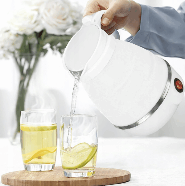 Bílá elektrická konvice nalévající horkou vodu do dvou sklenic s citronovou vodou na dřevěné podložce, v pozadí váza s bílými růžemi.