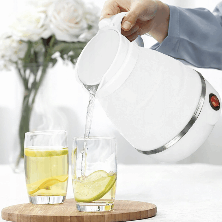 Bílá elektrická konvice nalévající horkou vodu do dvou sklenic s citronovou vodou na dřevěné podložce, v pozadí váza s bílými růžemi.