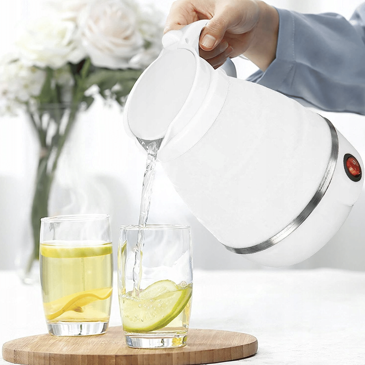 Bílá elektrická konvice nalévající horkou vodu do dvou sklenic s citronovou vodou na dřevěné podložce, v pozadí váza s bílými růžemi.