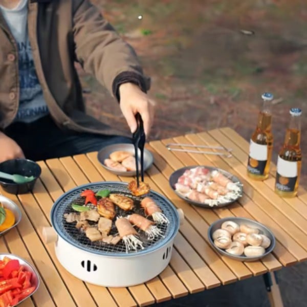 Přenosný gril s různými druhy masa a mořských plodů na dřevěném stole, v pozadí láhve piva a čerstvá zelenina.