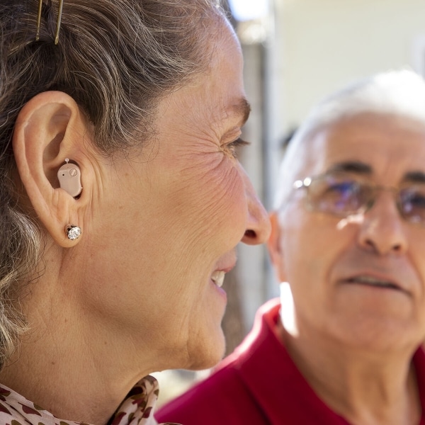 Starší žena s naslouchátkem v uchu a náušnicí se usmívá na rozmazanou postavu seniora v červeném oblečení v pozadí.