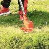 Sekání trávy pomocí elektrické strunové sekačky v oranžové barvě, zachycené v pohybu na zelené trávě s několika padlými listy.