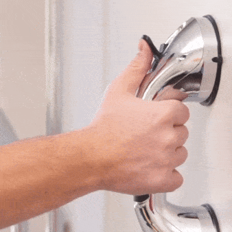 Ruka držící chromovanou sprchovou hlavici s kovovým uchycením na bílé koupelnové stěně.