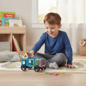 Malý chlapec v modrém tričku si hraje s barevnými magnetickými stavebnicovými kostkami ve tvaru vagónu na podlaze dětského pokoje.