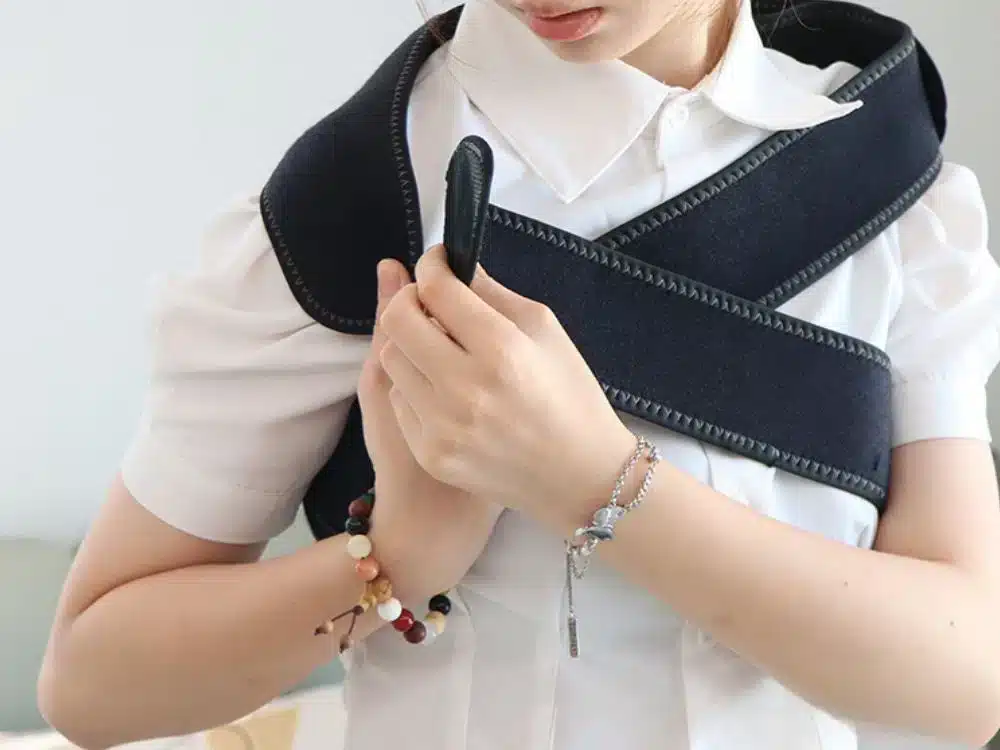 Žena v bílé košili s černou textilní ortézou nebo podporou na rameno, s barevným náramkem na zápěstí.