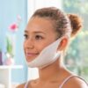 Žena s upraveným účesem v drdolu s bílou pleťovou maskou přes dolní část obličeje, sedící u okna v rozmazaném pozadí.