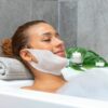 Žena odpočívající v lázni s obličejovou maskou, v pozadí svíčky a zelené listy navozující atmosféru relaxace a péče o pleť.