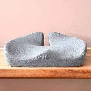 Šedý ergonomický sedací polštář s měkkým, zvlněným tvarem položený na dřevěné podlaze s růžovým pozadím.