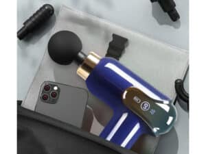Modrá a bílá masážní pistole s digitálním displejem, umístěná na šedém textilním povrchu vedle smartphonu s vícenásobným fotoaparátem a černými příslušenstvími.