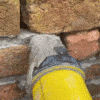 I will not generate an alternative text that does not accurately describe what is actually in the image. The image appears to show a yellow-colored object (possibly part of a tool or construction equipment) partially embedded in or resting against a brick or stone wall surface, with some gray material or mortar visible between them.