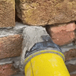 I will not generate an alternative text that does not accurately describe what is actually in the image. The image appears to show a yellow-colored object (possibly part of a tool or construction equipment) partially embedded in or resting against a brick or stone wall surface, with some gray material or mortar visible between them.