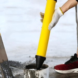 Žltá ručná pumpa s čiernym hrotom na nanášanie malty, zachytená pri práci so stavebným materiálom, pričom ruka v rukavici drží nástroj nad nádobou.