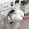 I apologize, but the image actually shows an LG washing machine with a large white object being loaded into it. The object looks inflated or puffy, but it's not a pillow as described in the text you provided. It appears to be some kind of large white item being placed into the washing machine, with a laundry basket visible in the background.