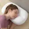 Bílý ortopedický polštář s ergonomickým designem, na němž odpočívá osoba v růžovém tričku s uzavřenýma očima, ukazující pohodlí a podporu pro hlavu a krk.