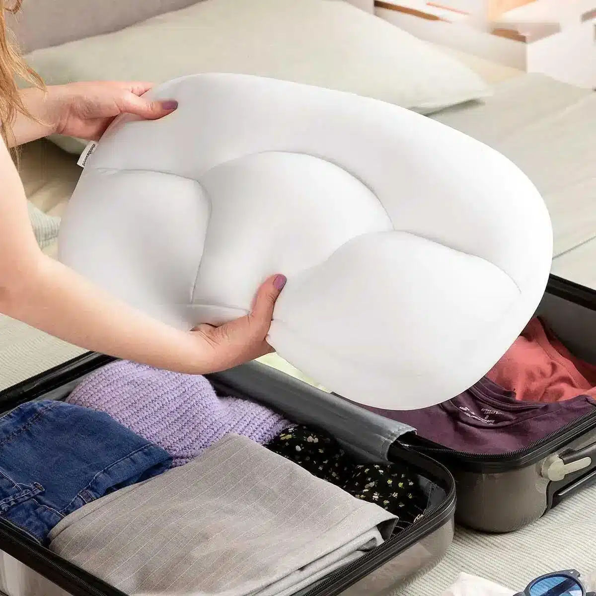 Bílý ergonomický Cloud polštář, který je držen rukama a nachází se částečně v otevřené cestovní tašce mezi oblečením a dalšími kusy výbavy.