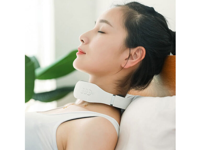 Žena s černými vlasy odpočívá s bílým krčním masážním přístrojem, oči má zavřené a vypadá uvolněně v minimalistickém prostředí s rostlinou v pozadí.