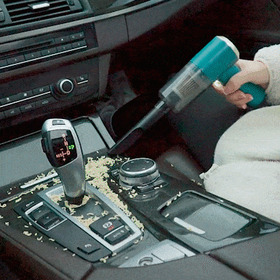 Ruční vysavač odstraňující drobky z interiéru automobilu, zaměřený na oblast řadicí páky a středové konzole.