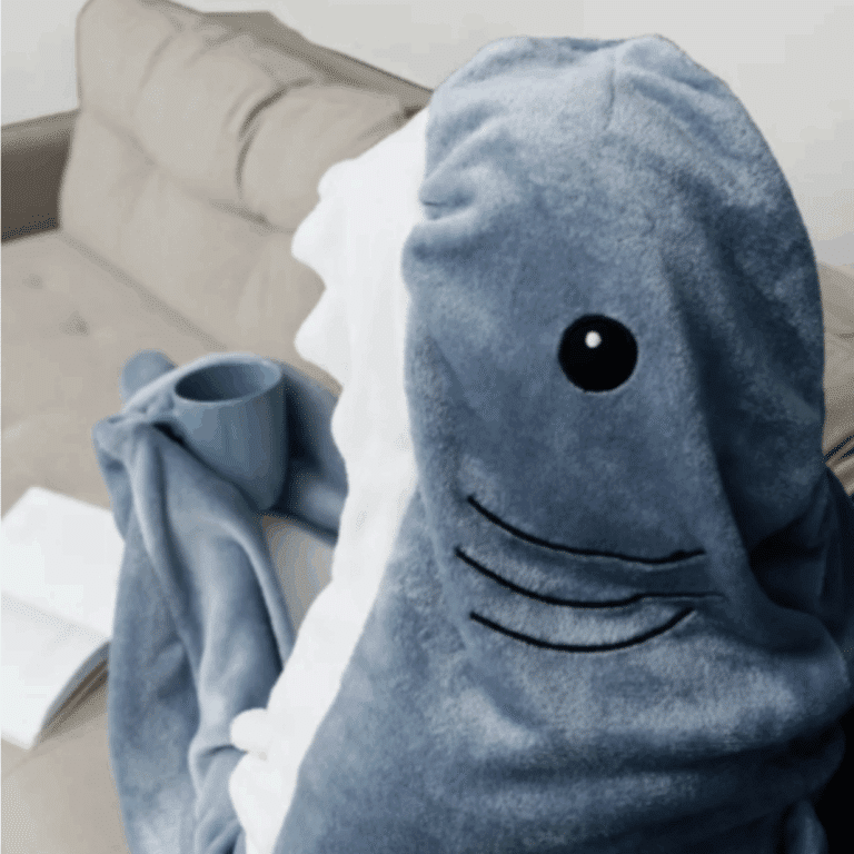 Modrá chlupatá deka ve tvaru žraloka s černýma očima a úsměvem, položená na gauči vedle zelené keramické šálky.