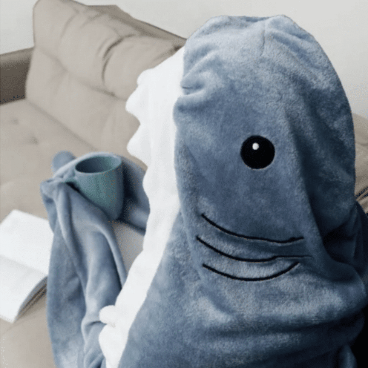 Modrá chlupatá deka ve tvaru žraloka s černýma očima a úsměvem, položená na gauči vedle zelené keramické šálky.