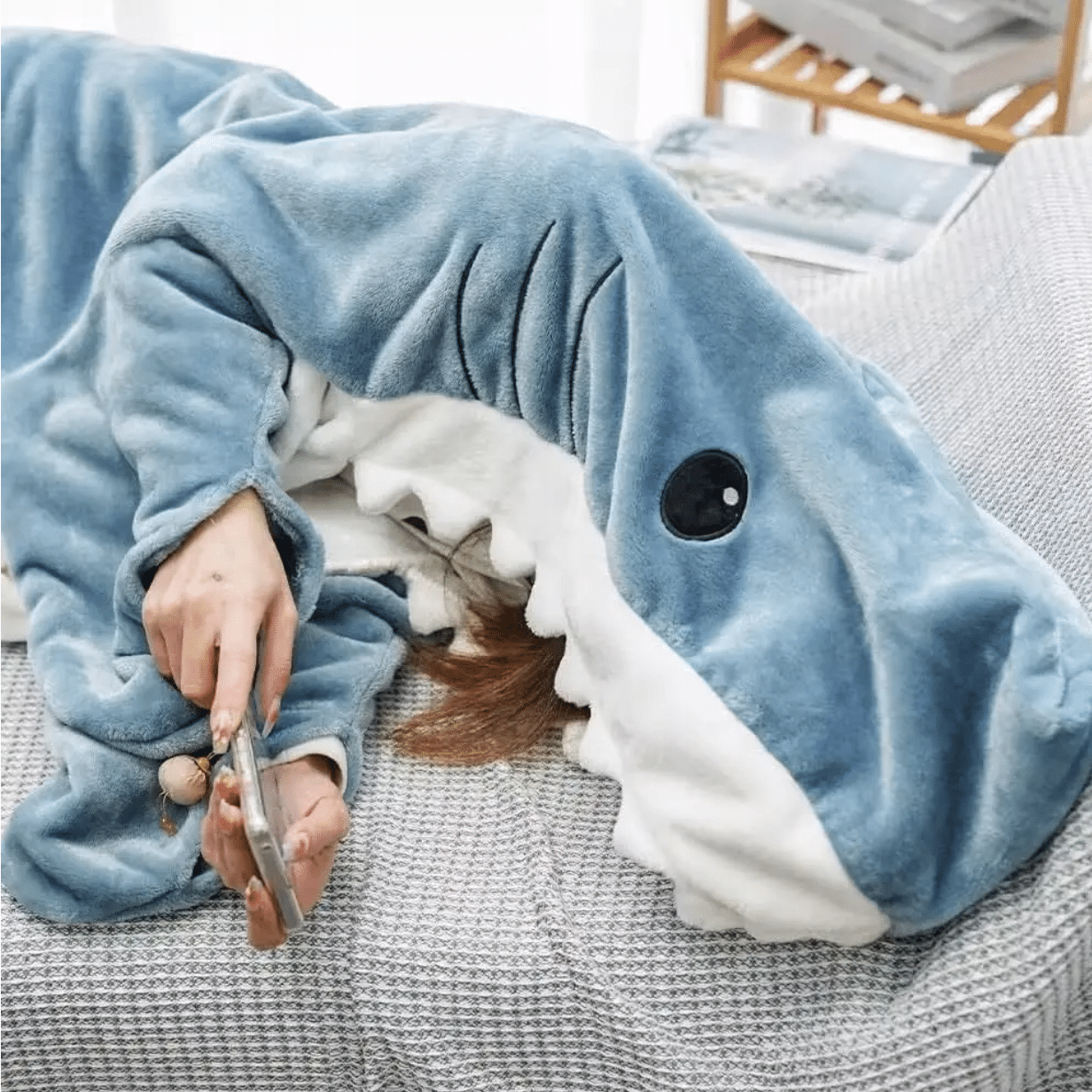 Plyšová deka ve tvaru žraloka, do které je zabalená osoba ležící na gauči a hrající si s mobilním telefonem.