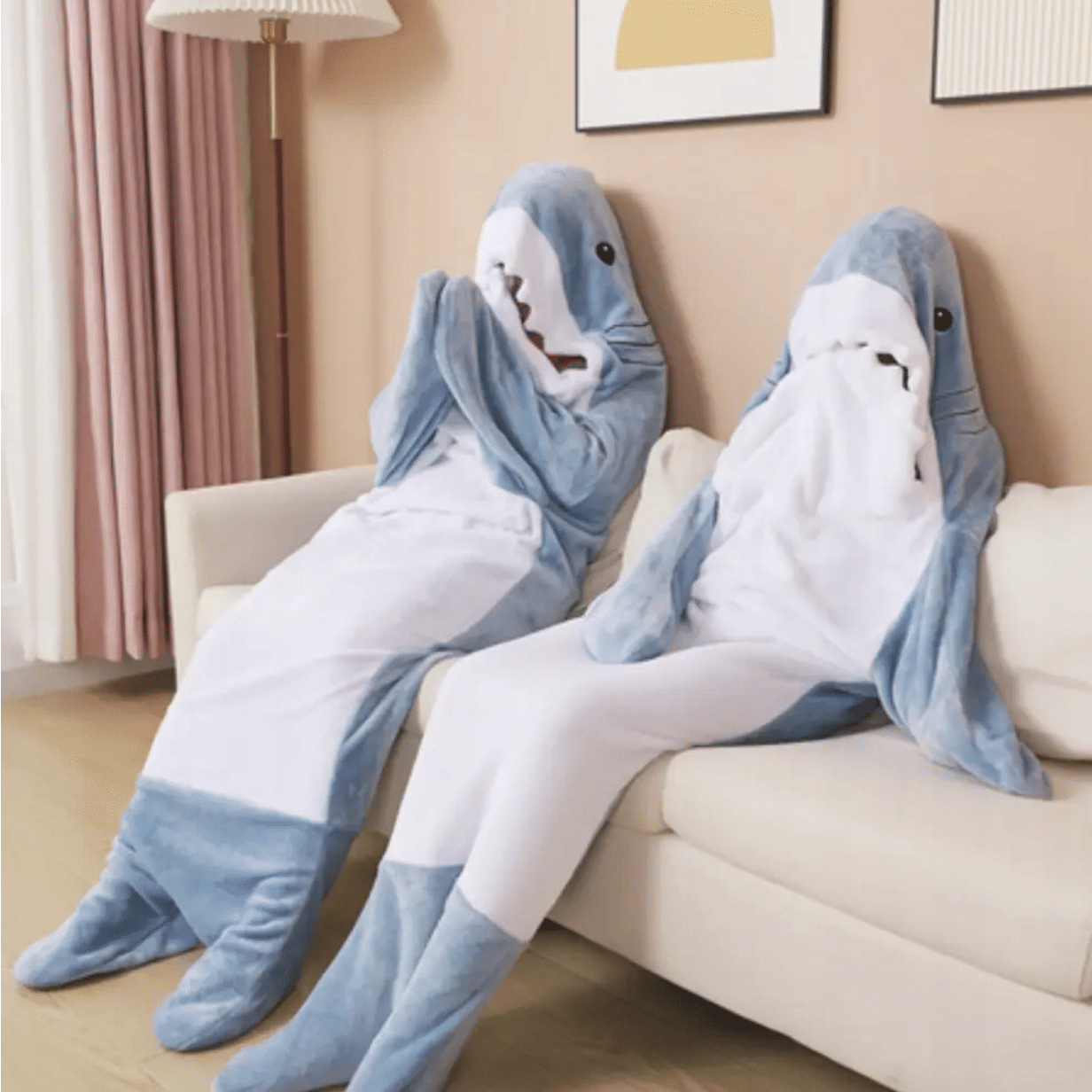 Dva plyšové žraloci v modrobílém provedení sedící vedle sebe na světlé pohovce v útulném pokoji s pastelovou výmalbou.