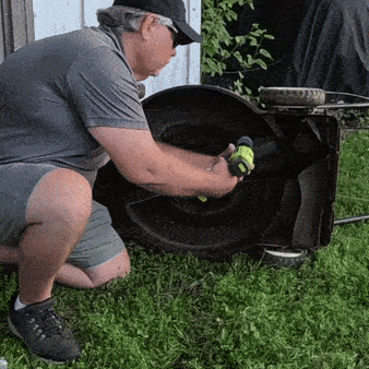 Muž ve věku v šedém tričku a čepici klečí na trávníku a otevírá černý venkovní gril připravený na grilování.