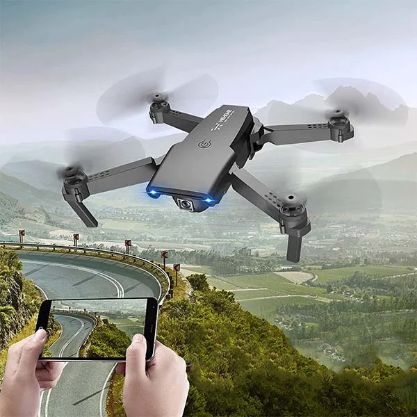 Kompaktný mini dron vo vzduchu nad horskou cestou, ovládaný rukami držiacimi diaľkový ovládač, s modro podsvietenými motormi a zabudovanou HD kamerou.