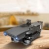 Kompaktný sivý dron so sklopenými ramenami vybavený HD kamerou, umiestnený na drevenej ploche s rozmazaným pozadím.