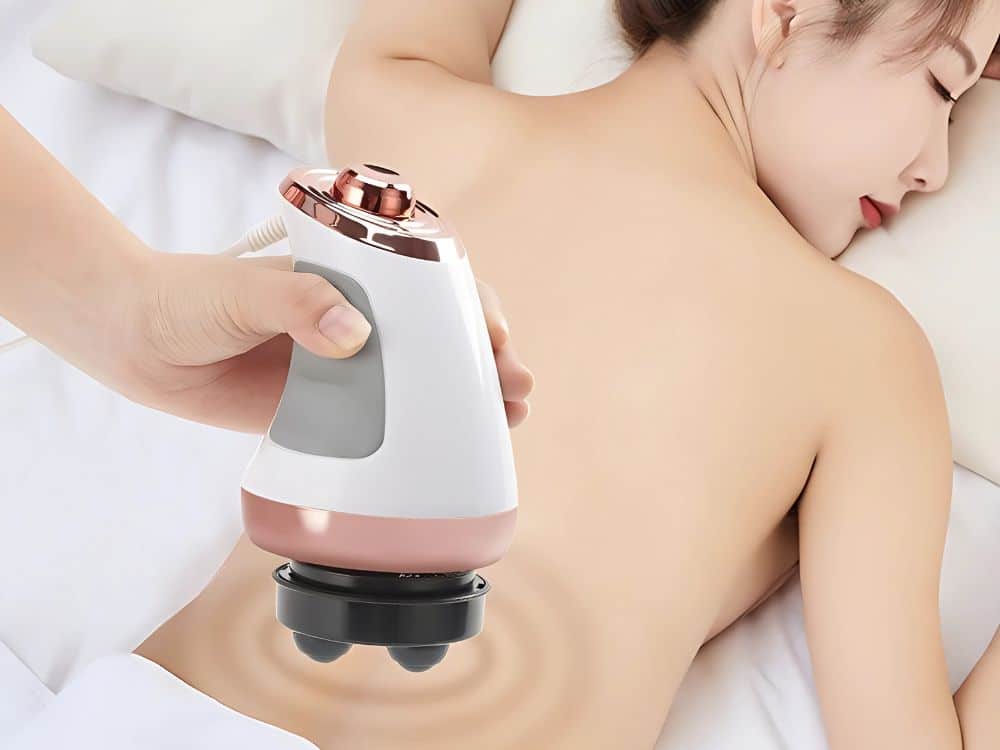 Masážní přístroj s ergonomickým designem bílo-růžové barvy, který je přiložen na záda odpočívající osoby na bílém lůžku.